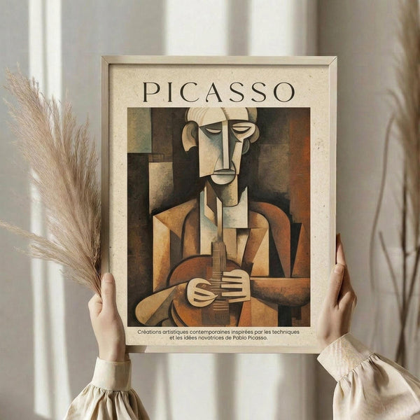 May include: Person in cream clothing holding framed Picasso-style cubist artwork showing angular musician figure in brown, orange and beige tones playing guitar, with dried pampas grass visible to left, against neutral curtained background