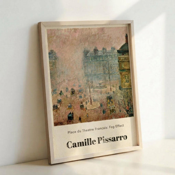 May include: Framed art print leaning against white wall showing Impressionist street scene with pink-toned sky, classical Parisian buildings, carriages and figures in square. Painting has soft, hazy atmosphere with muted colors. Light wood frame with white border displays artwork titled "Place du Theatre Francais: Fog Effect" and "Camille Pissarro" in black text below image.