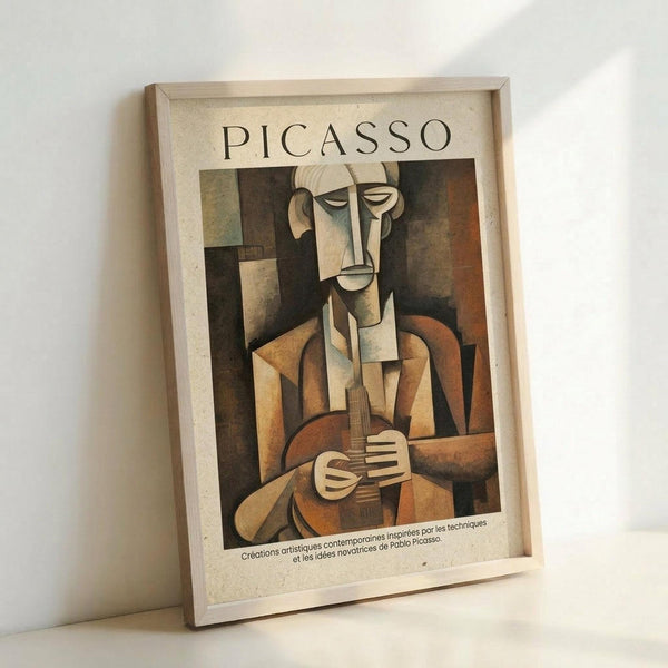 May include: Framed poster print of a cubist-style painting featuring an angular, geometric figure playing a guitar. The artwork displays earthy tones of brown, tan, and muted colors with the characteristic fragmented, abstract forms of early 20th century modernist art. The figure's face and body are depicted in geometric planes and shapes. French text appears at the bottom. The print is displayed in a light wood frame, leaning against a white wall on what appears to be a light-colored surface.