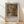 May include: Framed art print leaning against white wall on light wood floor with natural sunlight streaming across. Features impressionist-style painting of young woman with blonde hair wearing floral hat and coral-colored dress. Warm earth tones with soft brushstrokes. Text reads "PIERRE AUGUSTE exhibition" and "Young Girl with Hat" below image. Natural wood frame with cream matting.