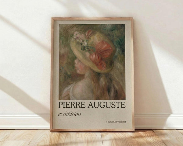 May include: Framed art print leaning against white wall on light wood floor with natural sunlight streaming across. Features impressionist-style painting of young woman with blonde hair wearing floral hat and coral-colored dress. Warm earth tones with soft brushstrokes. Text reads "PIERRE AUGUSTE exhibition" and "Young Girl with Hat" below image. Natural wood frame with cream matting.