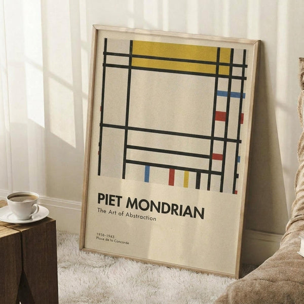 May include: A framed Mondrian-style abstract art poster with geometric black grid lines and blocks of yellow, red, and blue on cream background, leaning against white walls in a cozy corner with textured rug, wooden side table with white coffee cup, and beige knit blanket visible in natural lighting.