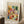 May include: Framed Paul Klee abstract watercolor poster featuring geometric squares and rectangles in vibrant oranges, greens, blues, purples, and earth tones. The artwork shows translucent overlapping shapes creating a mosaic-like composition. Light wooden frame leaning against white paneled wall on textured cream rug, with coffee cup on wooden block and beige fabric visible in cozy interior setting.