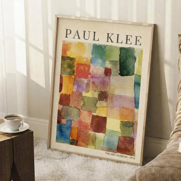 May include: Framed Paul Klee abstract watercolor poster featuring geometric squares and rectangles in vibrant oranges, greens, blues, purples, and earth tones. The artwork shows translucent overlapping shapes creating a mosaic-like composition. Light wooden frame leaning against white paneled wall on textured cream rug, with coffee cup on wooden block and beige fabric visible in cozy interior setting.