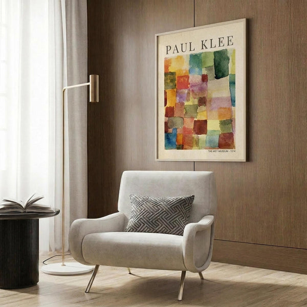May include: Framed Paul Klee abstract watercolor print with colorful geometric squares in blues, greens, oranges, and purples hanging on wood-paneled wall above cream upholstered armchair with geometric patterned pillow, next to brass floor lamp and dark round side table with open book