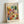 May include: Framed abstract watercolor art print with colorful geometric squares in warm oranges, yellows, greens, blues, and purples arranged in a mosaic pattern. The artwork features "PAUL KLEE" text at top and "THE MET MUSEUM - 1914" at bottom. Light wood frame leans against white wall corner on white surface, showing soft shadows.