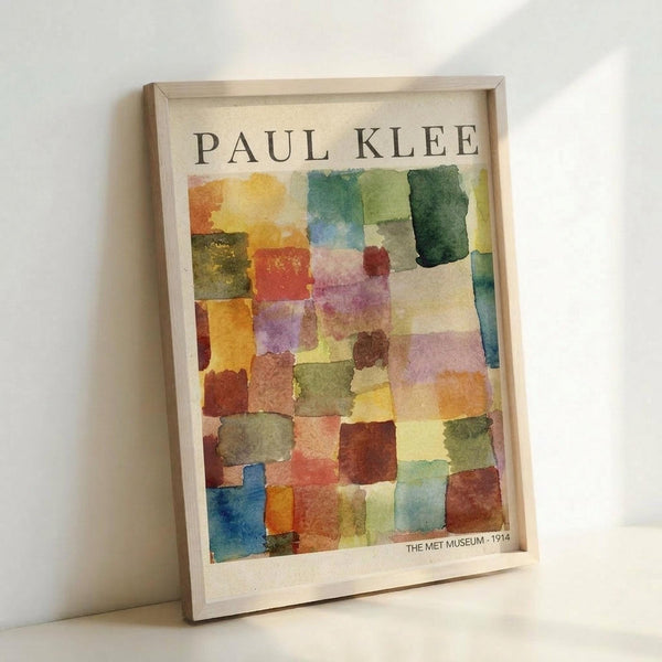 May include: Framed abstract watercolor art print with colorful geometric squares in warm oranges, yellows, greens, blues, and purples arranged in a mosaic pattern. The artwork features "PAUL KLEE" text at top and "THE MET MUSEUM - 1914" at bottom. Light wood frame leans against white wall corner on white surface, showing soft shadows.