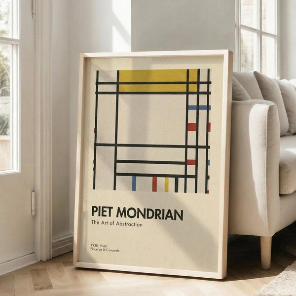 May include: Framed art print featuring Piet Mondrian's geometric abstract composition with black grid lines, yellow rectangle at top, small red and blue color blocks, and cream background. Text reads "PIET MONDRIAN The Art of Abstraction 1938-1943 Place de la Concorde" in black letters. Light wood frame leaning against white wall next to beige upholstered sofa in bright, airy room with tall windows.