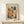 May include: Framed art print featuring Paul Klee's abstract watercolor composition with colorful geometric squares in warm oranges, cool blues, greens, and earth tones arranged in a mosaic pattern. Light wood frame displayed on white wooden surface with small wrapped gift box beside it against cream wall.