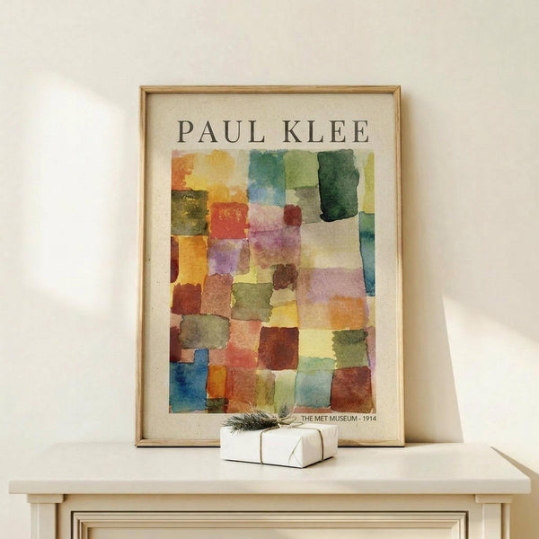 May include: Framed art print featuring Paul Klee's abstract watercolor composition with colorful geometric squares in warm oranges, cool blues, greens, and earth tones arranged in a mosaic pattern. Light wood frame displayed on white wooden surface with small wrapped gift box beside it against cream wall.