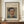 May include: Framed art print featuring a vintage portrait of a man with dark hair and mustache wearing a blue jacket with yellow collar details. The artwork has an impressionistic painting style with soft brushstrokes and muted colors. The print is displayed in a thin brown wooden frame on a wooden sideboard alongside a white ribbed ceramic vase and stacked books, against cream colored walls with white wainscoting.