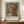 May include: Exhibition poster featuring impressionist-style painting of young woman with flowing blonde hair wearing coral-pink hat with flowers, rendered in soft brushstrokes with muted green background. Framed print displayed on wooden sideboard alongside white ceramic vase and stacked books in elegant room with cream walls and traditional molding.