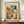 May include: Framed Paul Klee abstract watercolor print featuring mosaic-like squares in warm oranges, yellows, greens, blues, and purples arranged in a grid pattern. The cream-colored poster shows "PAUL KLEE" at top and "THE MET MUSEUM - 1914" at bottom, displayed in a thin brown wooden frame on a walnut credenza beside a white ceramic vase and stacked books against white wainscoting walls.