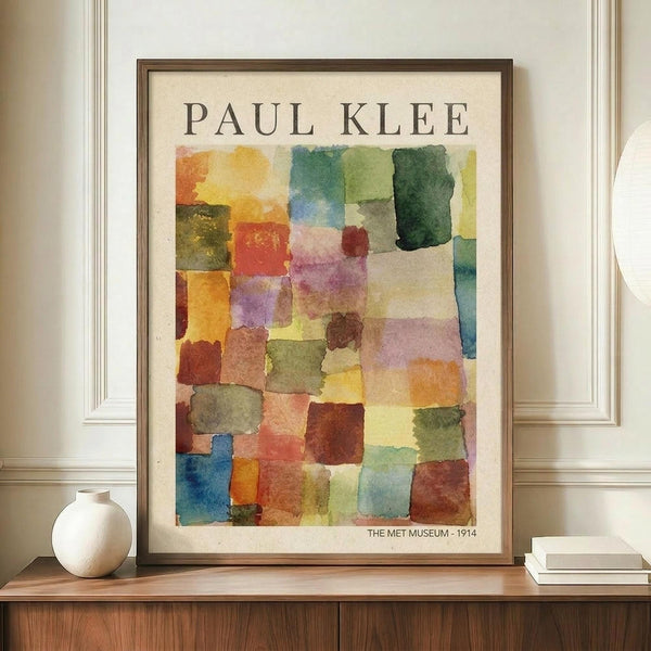 May include: Framed Paul Klee abstract watercolor print featuring mosaic-like squares in warm oranges, yellows, greens, blues, and purples arranged in a grid pattern. The cream-colored poster shows "PAUL KLEE" at top and "THE MET MUSEUM - 1914" at bottom, displayed in a thin brown wooden frame on a walnut credenza beside a white ceramic vase and stacked books against white wainscoting walls.