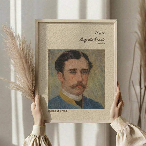 May include: Hands holding a framed reproduction of a Renoir portrait showing a man with dark hair and mustache wearing a blue military-style jacket with yellow trim. The beige textured mat displays the artist name and "portrait of a man" text. Dried wheat stalks visible on left side against soft curtained window background.