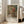 May include: Framed art print leaning against white wall next to beige upholstered chair in bright living room with large windows. Print features soft impressionist painting of young woman with flowing blonde hair wearing coral pink bow, rendered in warm peachy tones with green background. White mat displays "PIERRE AUGUSTE exhibition" text and artwork title below image.