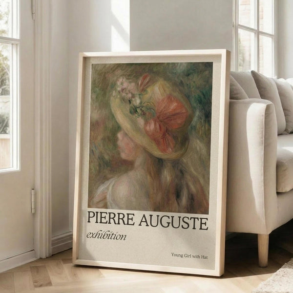 May include: Framed art print leaning against white wall next to beige upholstered chair in bright living room with large windows. Print features soft impressionist painting of young woman with flowing blonde hair wearing coral pink bow, rendered in warm peachy tones with green background. White mat displays "PIERRE AUGUSTE exhibition" text and artwork title below image.