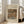 May include: Framed art print leaning against white wall next to beige armchair in bright room with large windows. Print features Renoir still life painting of oranges and bananas in warm earth tones on cream textured background with black text above and below artwork. Light wood frame, approximately 2-3 feet tall, positioned on light wooden floor beside furniture leg.