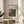 May include: Framed reproduction of Renoir's impressionist landscape painting showing the Peninsula of Saint-Jean with soft greens, browns, and blues depicting trees, figures, and water. The print is displayed in a thin wooden frame on a round wooden side table next to a beige upholstered chair with white textured pillow, near a plant and white curtains in natural sunlight.