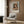 May include: Framed vintage-style poster featuring Renoir's impressionist painting of a young girl wearing a pink bonnet and brown clothing, displayed on wood-paneled wall above a beige upholstered armchair with geometric patterned pillow. Modern living room setting with brass floor lamp, dark round side table with open book, and natural lighting from window with sheer curtains.