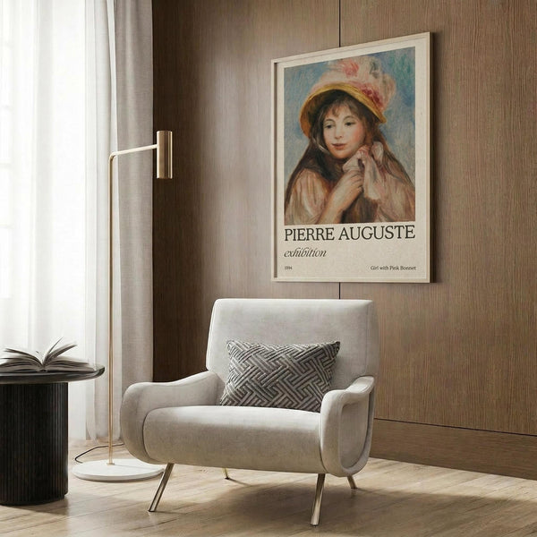 May include: Framed vintage-style poster featuring Renoir's impressionist painting of a young girl wearing a pink bonnet and brown clothing, displayed on wood-paneled wall above a beige upholstered armchair with geometric patterned pillow. Modern living room setting with brass floor lamp, dark round side table with open book, and natural lighting from window with sheer curtains.