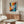 May include: Abstract expressionist artwork poster with explosive radiating design in vibrant blues, oranges, and yellows emanating from central white circle with red dot. Framed print displayed above mid-century walnut credenza in modern living room with white curved chair, glass coffee table, and minimalist brass floor lamp against neutral walls.