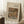 May include: Framed art print leaning against white wall corner on textured cream rug. Print shows Auguste Renoir still life painting of oranges and bananas in warm golden tones with beige matting and thin wood frame. Coffee cup on wooden block visible nearby with soft natural lighting creating window shadows.