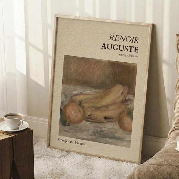 May include: Framed art print leaning against white wall corner on textured cream rug. Print shows Auguste Renoir still life painting of oranges and bananas in warm golden tones with beige matting and thin wood frame. Coffee cup on wooden block visible nearby with soft natural lighting creating window shadows.