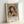 May include: Framed art print leaning against white wall showing Renoir's impressionist painting of a young woman wearing a yellow and white bonnet with pink flowers. The portrait features soft, warm tones with the subject's hands positioned near her face. Below the artwork is exhibition-style text reading "PIERRE AUGUSTE exhibition" with "Girl with Pink Bonnet 1894" in smaller text. The print is displayed in a light wooden frame.