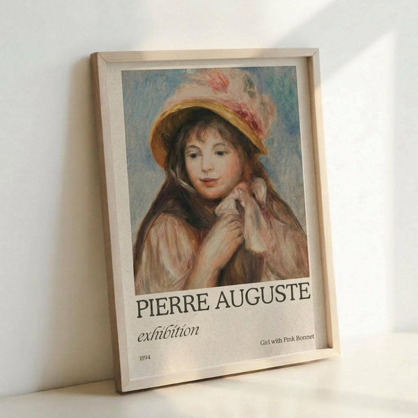 May include: Framed art print leaning against white wall showing Renoir's impressionist painting of a young woman wearing a yellow and white bonnet with pink flowers. The portrait features soft, warm tones with the subject's hands positioned near her face. Below the artwork is exhibition-style text reading "PIERRE AUGUSTE exhibition" with "Girl with Pink Bonnet 1894" in smaller text. The print is displayed in a light wooden frame.