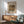 May include: Framed Renoir poster featuring oranges and bananas still life painting in warm tones, displayed above a mid-century wooden sideboard with white table lamp, next to sage green upholstered armchair with wood frame in sunlit modern living room with white walls.