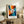 May include: Framed abstract expressionist poster with explosive radiating design in vibrant orange, blue, red and black paint splatters emanating from central white circle, displayed on rustic wooden sideboard alongside fern plant, ceramic vase, and stacked books in neutral-toned living room setting
