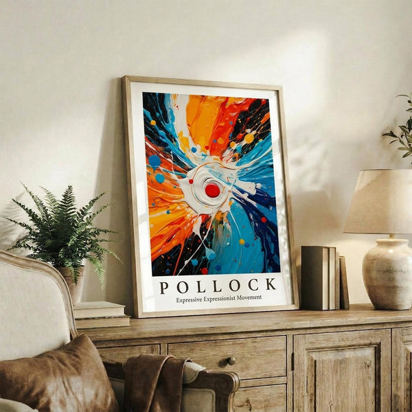 May include: Framed abstract expressionist poster with explosive radiating design in vibrant orange, blue, red and black paint splatters emanating from central white circle, displayed on rustic wooden sideboard alongside fern plant, ceramic vase, and stacked books in neutral-toned living room setting