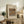 May include: Framed art print featuring Renoir's still life painting of oranges and bananas in warm earth tones, displayed on a rustic wooden sideboard alongside potted fern, ceramic vase, stacked books, and brown leather chair in neutral-toned living room setting.