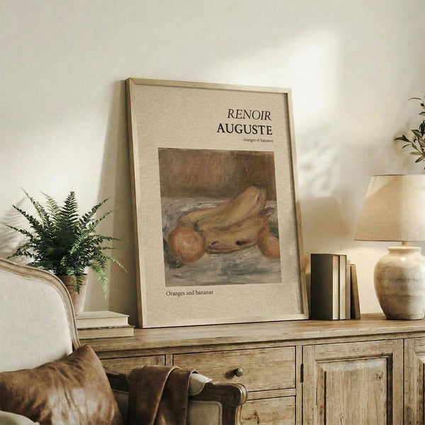May include: Framed art print featuring Renoir's still life painting of oranges and bananas in warm earth tones, displayed on a rustic wooden sideboard alongside potted fern, ceramic vase, stacked books, and brown leather chair in neutral-toned living room setting.