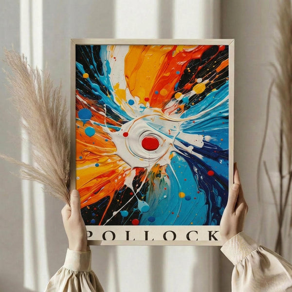 May include: Person holding framed abstract art print featuring explosive burst pattern radiating from central white eye with red center. Vibrant orange, blue, black paint splatters and streaks create dynamic movement. Light wooden frame, neutral background with decorative dried grass stems visible on left side.