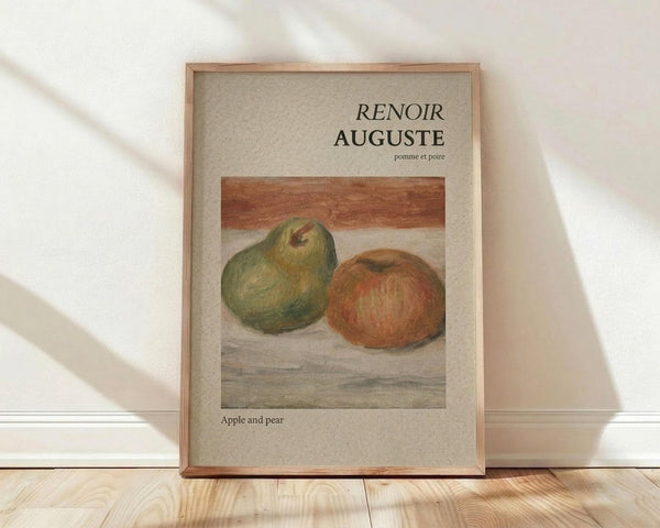 May include: Framed art print featuring Auguste Renoir's still life painting of a green pear and red apple on neutral background. Print has beige matting with black text reading "RENOIR AUGUSTE" and "pomme et poire" above the artwork, "Apple and pear" below. Natural wood frame leans against white wall on light wooden floor with soft window light shadows.