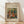 May include: Framed art print of Renoir's impressionist painting showing women and children in a pastoral outdoor scene with warm earth tones and soft brushstrokes, displayed in thin wooden frame against white wall on light wooden floor with natural window lighting creating diagonal shadows