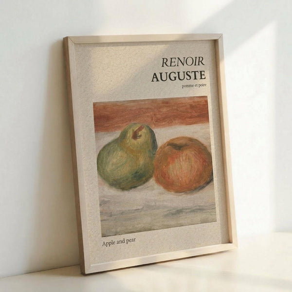 May include: Framed art print leaning against white wall showing Renoir's still life painting of a green pear and red apple on wooden surface. Light wooden frame with cream matting, soft natural lighting creating gentle shadows on white background.