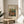 May include: Framed art print of Renoir's colorful Impressionist painting showing figures in a garden scene with warm reds, greens, and earth tones, displayed on a round wooden side table next to a beige upholstered chair with natural sunlight casting shadows across white walls and green plant leaves visible in corner of bright, minimalist room setting.