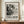 May include: Framed black and white abstract art poster with splattered paint design mounted on cream wall above wooden sideboard. White ribbed ceramic vase and stack of books sit beside the brown wooden frame. The poster features bold text reading "POLLOCK" and exhibition details with dates. Abstract expressionist style artwork shows dynamic paint splatters and organic forms in monochrome palette.