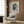 May include: Black and white abstract Pollock exhibition poster with splattered paint design mounted on wood-paneled wall above beige upholstered armchair with geometric patterned pillow. Modern room setting features gold floor lamp, dark round side table with open book, and neutral curtains. Poster shows chaotic paint splatters with bold text below.
