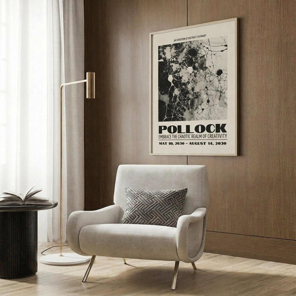 May include: Black and white abstract Pollock exhibition poster with splattered paint design mounted on wood-paneled wall above beige upholstered armchair with geometric patterned pillow. Modern room setting features gold floor lamp, dark round side table with open book, and neutral curtains. Poster shows chaotic paint splatters with bold text below.