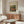 May include: Framed art print featuring Impressionist painting of two figures in warm oranges, greens and blues displayed above a mid-century modern wooden credenza with sliding doors. White sculptural chair and glass-top oval coffee table visible in foreground. Minimalist room with neutral walls and natural lighting from window.