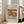 May include: Framed art print leaning against beige sofa in bright living room with large windows. Print shows Renoir still life painting of green pear and red apple on white surface. Light wood frame, cream matting, natural sunlight streaming across wooden floors.
