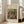 May include: Framed poster print of a Renoir impressionist painting showing figures in an outdoor garden setting with soft brushstrokes in warm oranges, greens, and blues. The artwork is displayed in a light wooden frame against a textured beige mat, leaning against a white wall next to a cream-colored armchair near tall windows with natural lighting.