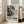 May include: A framed abstract art exhibition poster featuring black and white paint splatters and drips in Jackson Pollock's signature style. The poster displays "POLLOCK" in bold letters with "EMBRACE THE CHAOTIC REALM OF CREATIVITY" and exhibition dates "MAY 18, 2030 - AUGUST 14, 2030". The light wood frame leans against a white wall next to a beige sectional sofa in a bright living room with tall windows and hardwood floors.