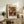 May include: Framed art print of Renoir's 1919 floral painting featuring vibrant orange, red and white chrysanthemums in a blue and white ceramic vase, displayed on rustic wooden sideboard alongside fern plant, ceramic pottery, books and table lamp in warm neutral living room setting