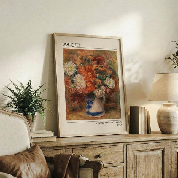 May include: Framed art print of Renoir's 1919 floral painting featuring vibrant orange, red and white chrysanthemums in a blue and white ceramic vase, displayed on rustic wooden sideboard alongside fern plant, ceramic pottery, books and table lamp in warm neutral living room setting