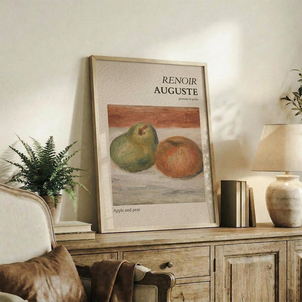 May include: Framed art print featuring Auguste Renoir's still life painting of green pears and a red apple on neutral background. Print displayed on rustic wooden sideboard with cream upholstered chair, potted fern, ceramic vase, and stacked books in warm-toned living room setting.